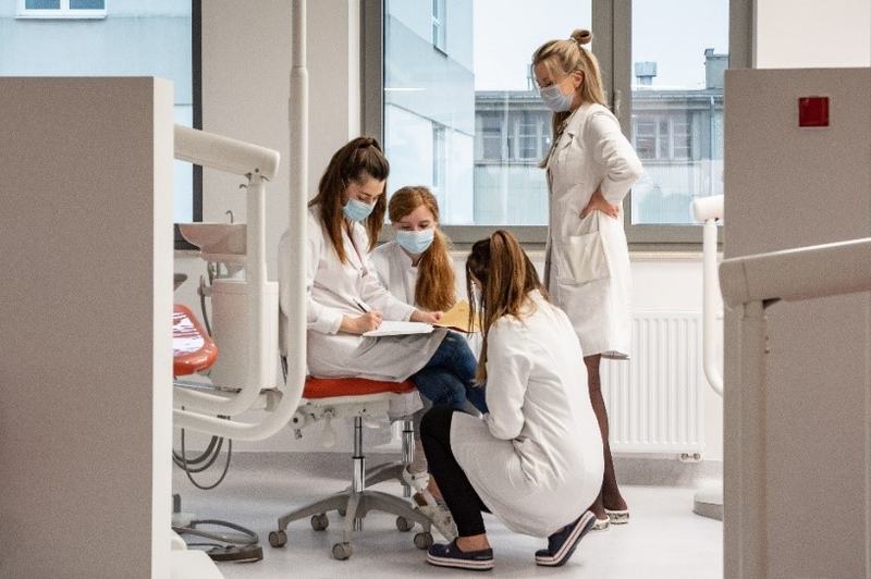 Cztery studentki stomatologii w białych fartuchach i maseczkach medycznych podczas zajęć w gabinecie dentystycznym