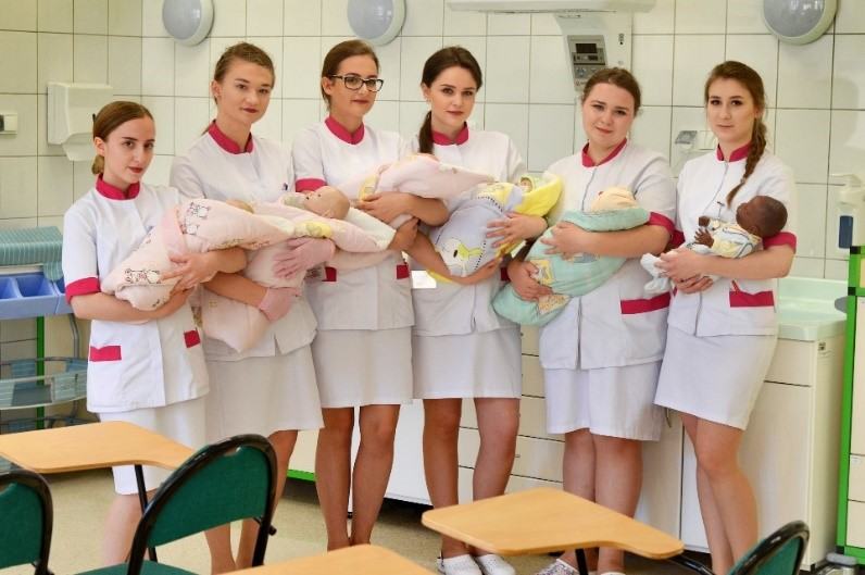 ześć studentek położnictwa w białych fartuchach trzymających na rękach lalki do ćwiczeń imitujące noworodki w sali dydaktycznej