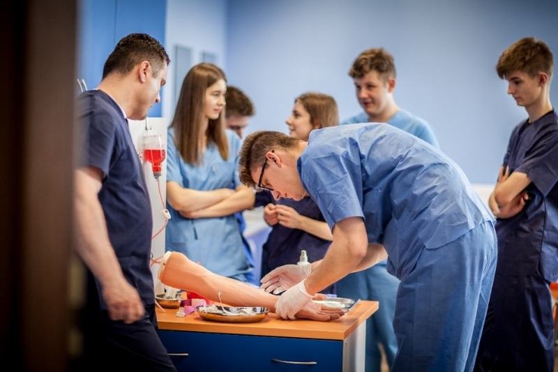 Student medycyny w rękawiczkach ćwiczący pobieranie krwi na sztucznym modelu ręki pod okiem grupy i instruktora